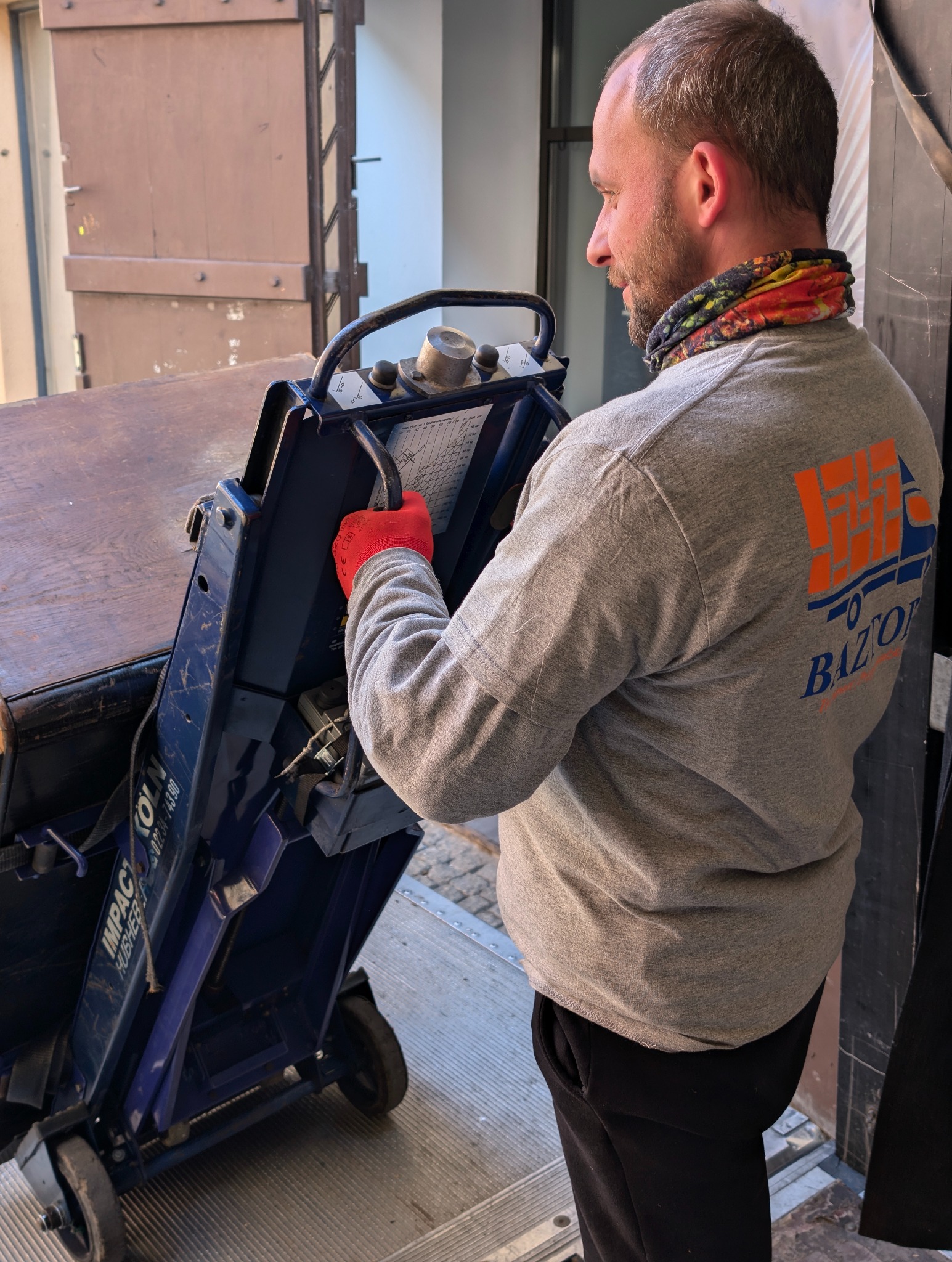 Pracownik w szarej bluzie z logo firmy przeprowadzkowej Baztor obsługuje wózek schodowy Impact Koln podczas transportu mebla, widoczne czerwone rękawice ochronne, tło stanowi brama.