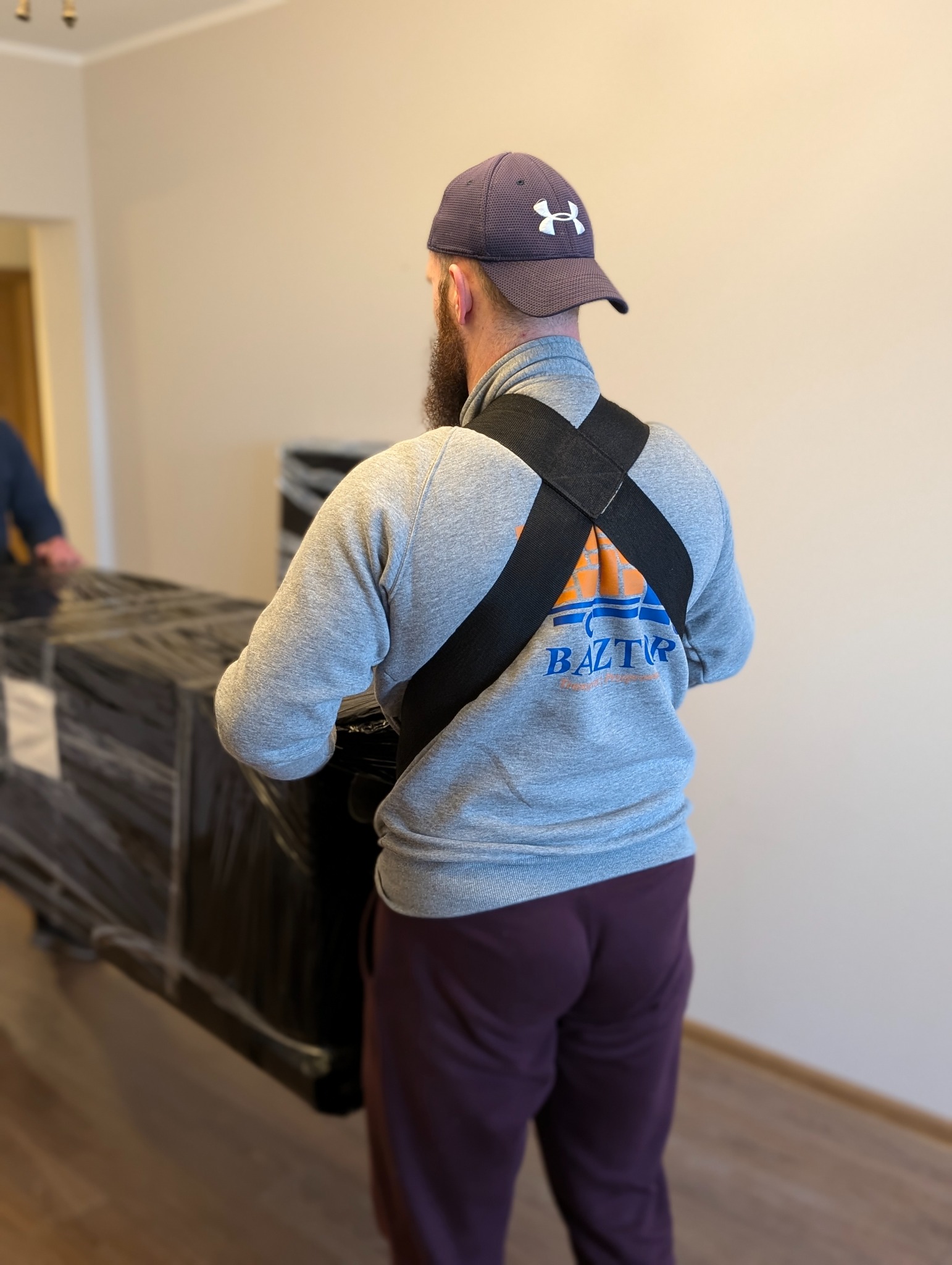 Pracownik firmy przeprowadzkowej z brodą w czapce z daszkiem, ubrany w szarą bluzę z logo firmy 'BAZTUR' na plecach, przenosi zapakowany w folię stretch mebel za pomocą specjalnych pasów nośnych.