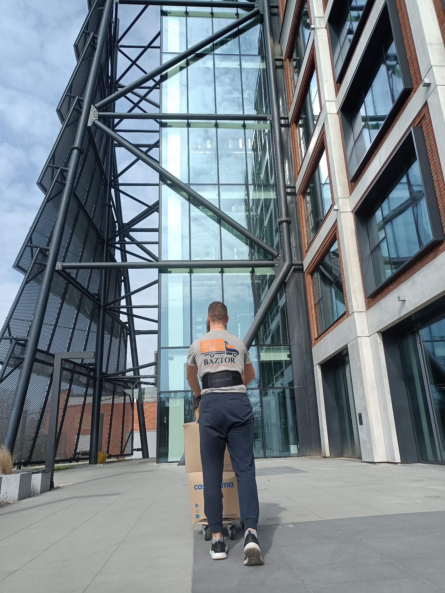 Pracownik firmy przeprowadzkowej w szarej koszulce z logo BAZTOR, widoczny od tyłu, ciągnie wózek z kartonami na tle nowoczesnej architektury biurowej z metalową konstrukcją i szklaną fasadą.