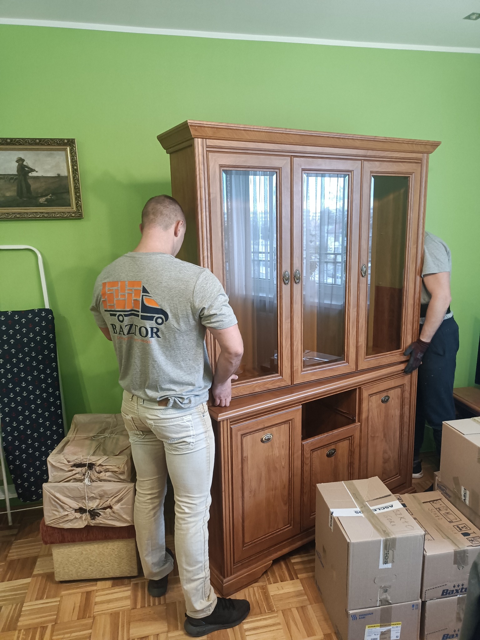 Dwóch mężczyzn w trakcie przenoszenia drewnianego kredensu z przeszklonymi drzwiami; jeden z nich ma na sobie szarą koszulkę z logo firmy transportowej 'BAZIOR', a obok stoją zapakowane kartony...