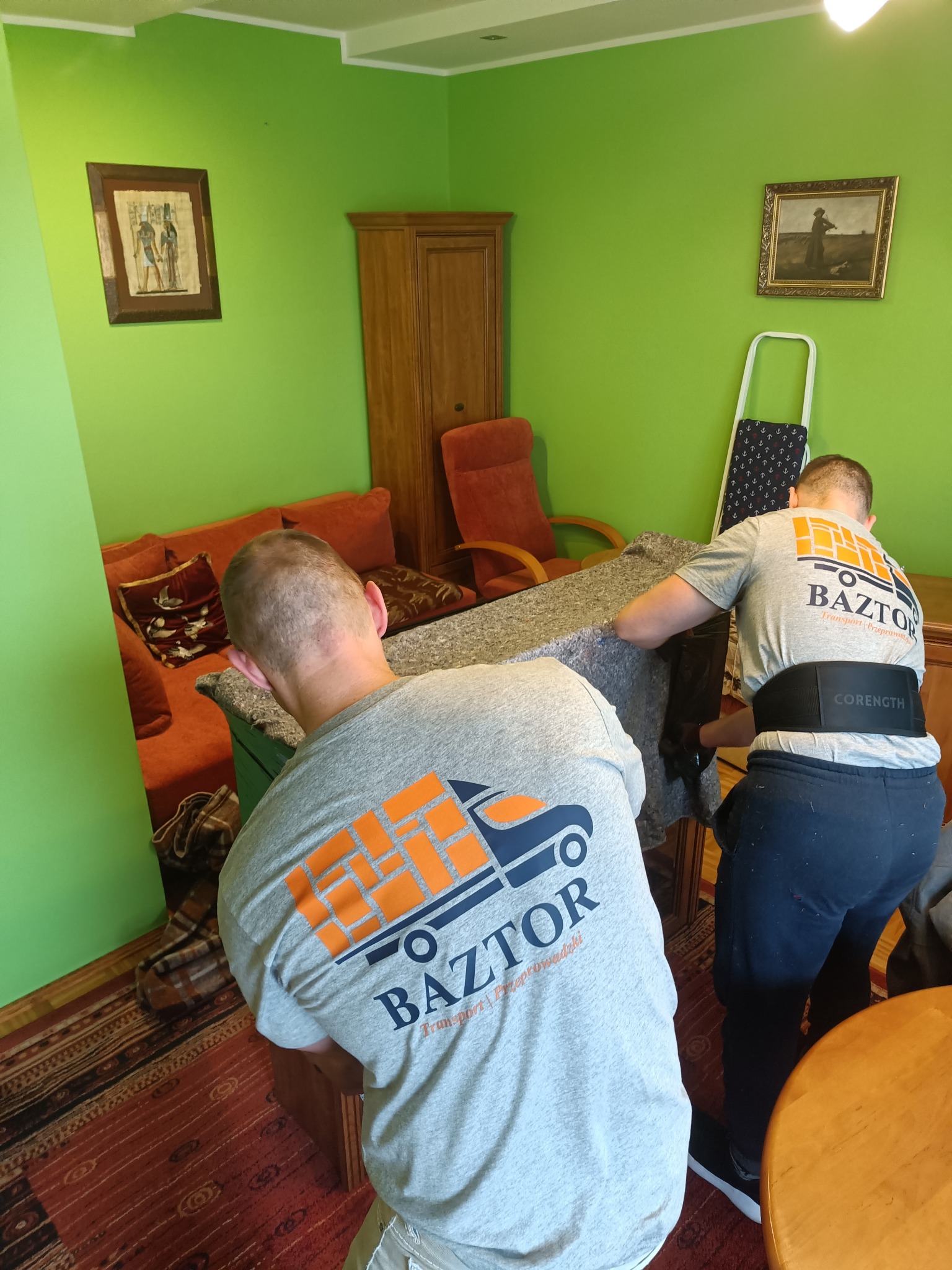 Dwóch mężczyzn w szarych koszulkach z logo firmy transportowej Baztor przenosi mebel owinięty w szary materiał w zielonym pokoju z dywanem o wzorzystym deseniu.