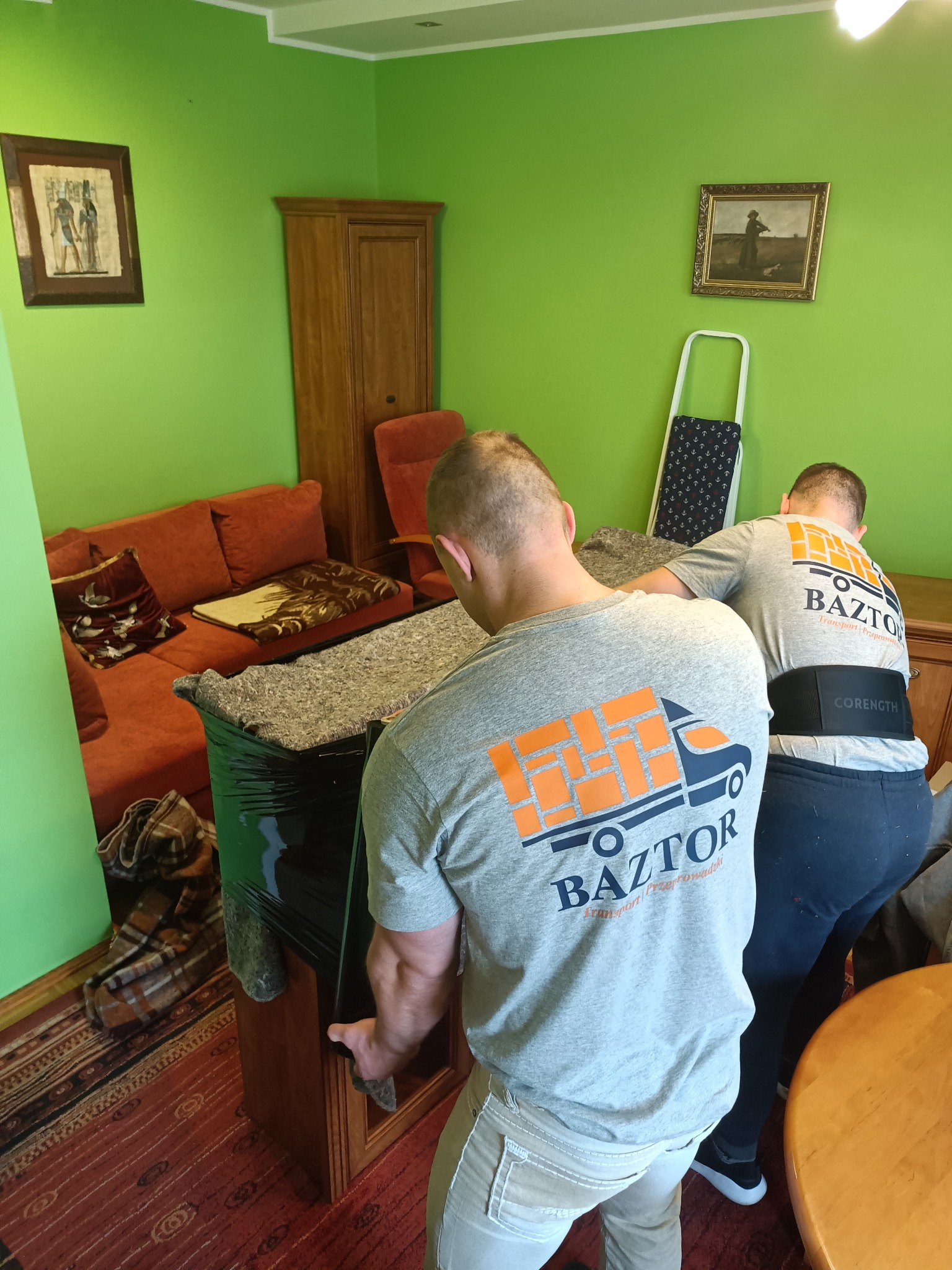 Dwóch mężczyzn w szarych koszulkach z logo firmy transportowej BAZTOR przenosi owiniętą w folię i koc szafkę w zielonym pokoju z obrazami na ścianach.
