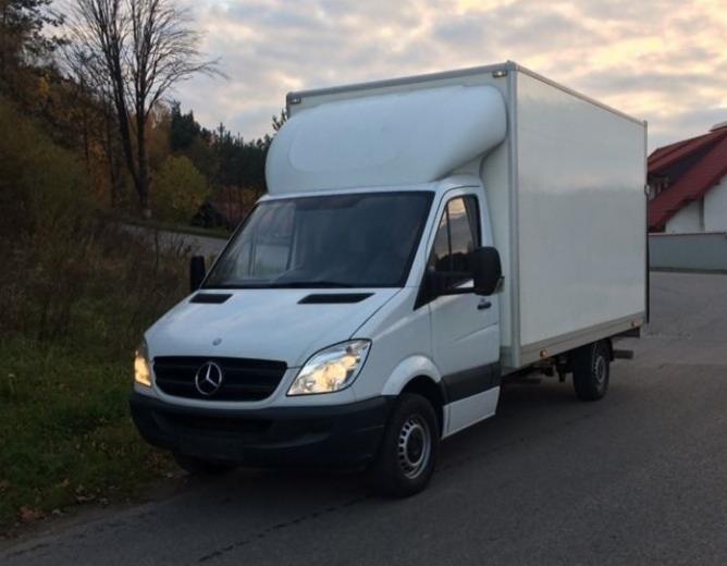 Biały samochód dostawczy Mercedes-Benz Sprinter z zabudową kontenerową, zaparkowany na poboczu drogi, widok z przodu i boku, jesienne drzewa w tle.