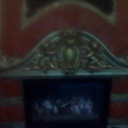 Usługi ogulno buwldowlane - Częściowo widoczny kominek z dekoracyjnym, złoconym nadprożem z ornamentami i obrazem w ciemnej obudowie, całość na tle czerwonej ściany.