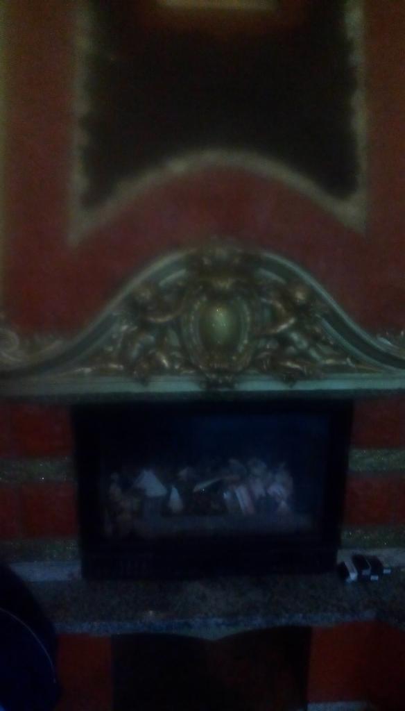 Częściowo widoczny kominek z dekoracyjnym, złoconym nadprożem z ornamentami i obrazem w ciemnej obudowie, całość na tle czerwonej ściany.