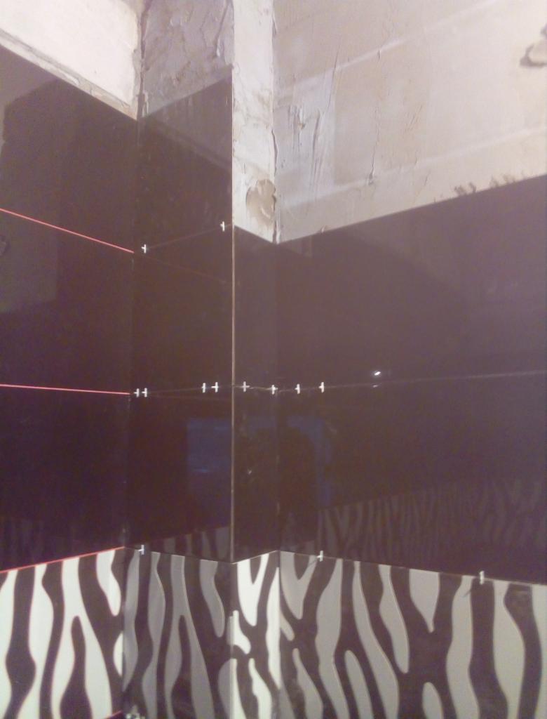 Układanie ciemnych płytek w łazience z dolnym rzędem płytek w zebrę, widoczny laserowy niwelator i krzyżyki dystansowe, surowy stan ściany nad płytkami.