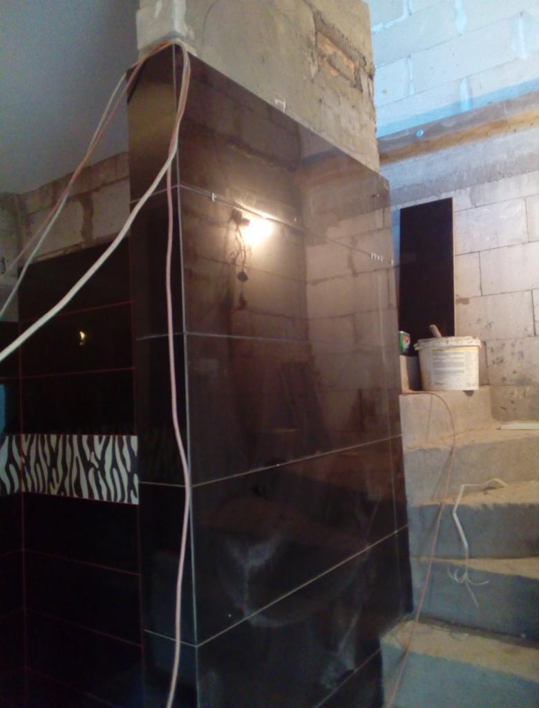 Częściowo wykończona ściana z czarnych, błyszczących płytek w łazience, z fragmentem dekoracyjnej mozaiki w zebrę, widoczne kable elektryczne i surowe ściany z betonu komórkowego oraz schody.