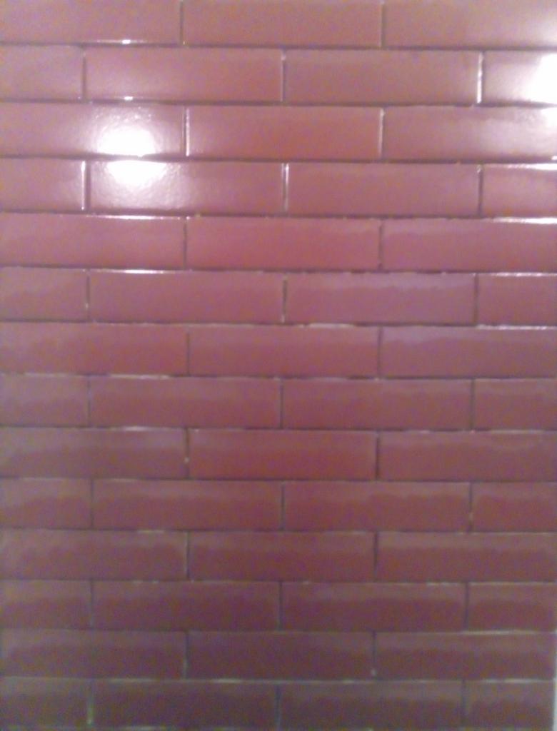 Ściana wyłożona błyszczącymi, bordowymi płytkami w kształcie cegiełek, ułożonych poziomo z przesunięciem.