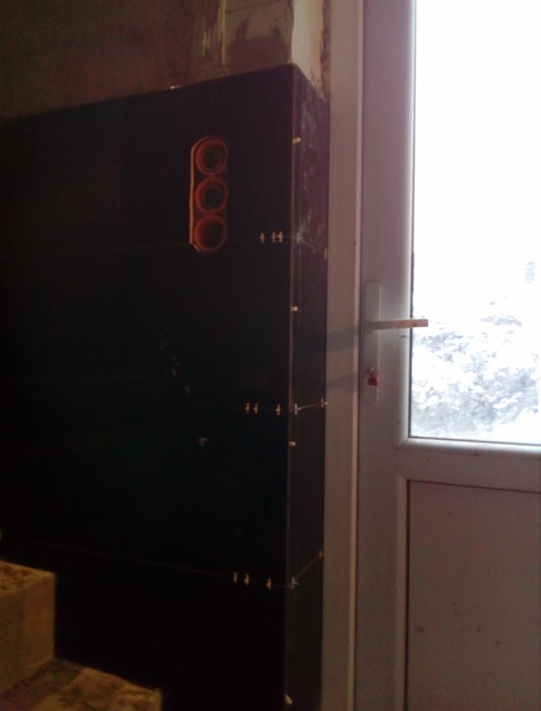 Ciemna ściana z wyciętymi otworami na rury, przytwierdzona do ściany za pomocą sznurków i kołków dystansowych, obok białych drzwi balkonowych.