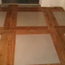 Nowa podłoga w łazience z jasnych, kwadratowych płytek otoczonych drewnopodobnymi panelami tworzącymi krzyżowy wzór.