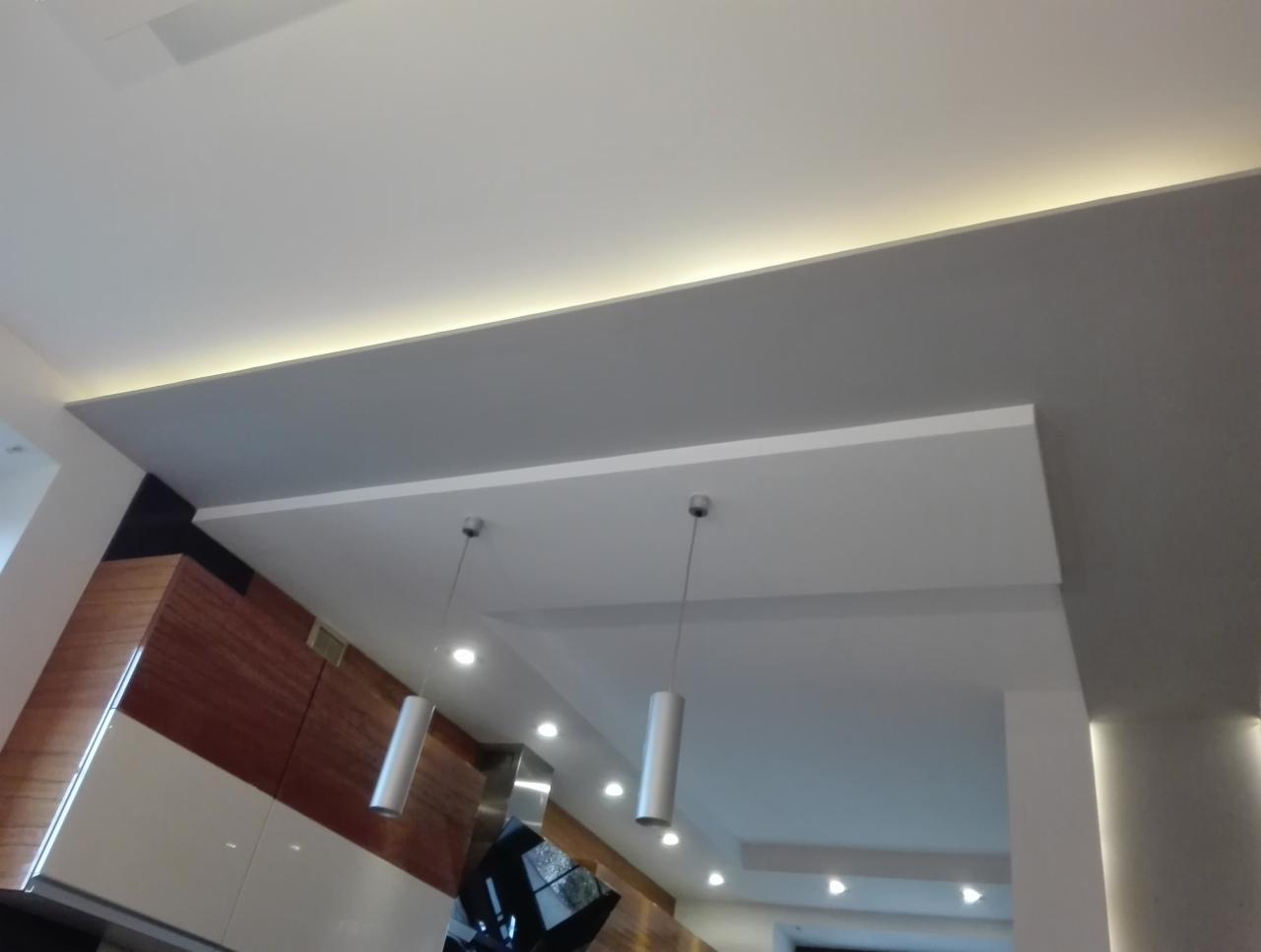 Nowoczesny sufit podwieszany z oświetleniem LED wzdłuż krawędzi, cylindryczne lampy wiszące, fragment zabudowy kuchennej z fornirem drewnopodobnym.