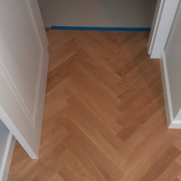 MB Constructions Michał Borkowski - Ułożona parkietowa podłoga w jodełkę, widok z góry na fragment pomieszczenia z otwartymi białymi drzwiami i niepomalowaną ścianą zabezpieczoną niebieską taśmą malarską.