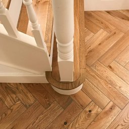 MB Constructions Michał Borkowski - Ułożona w jodełkę parkietowa podłoga z dębowego drewna, z widocznym fragmentem białej balustrady schodów i jej okrągłą podstawą.