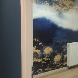 MB Constructions Michał Borkowski - Ściana z tapetą w odcieniach błękitu i złota, widoczna przez futrynę drzwi, z grzejnikiem na tle i jasną podłogą.