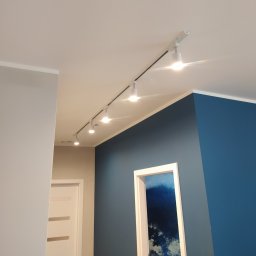 MB Constructions Michał Borkowski - Wnętrze z niebieską ścianą, białymi drzwiami z poziomymi przeszkleniami i szynoprzewodem z reflektorami punktowymi na suficie.