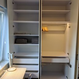 MB Constructions Michał Borkowski - Wnętrze białej szafy wnękowej z oświetleniem LED, półkami i szufladami, widoczne również wieszaki i złożone ręczniki, obok biurko z lampką i krzesłem.