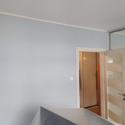 MB Constructions Michał Borkowski - Świeżo pomalowane na jasnoniebiesko ściany pokoju, zabezpieczone meble folią malarską, widoczny fragment drzwi z ościeżnicą i włącznik światła.