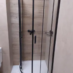 Narożna kabina prysznicowa z czarnymi profilami i armaturą, zamontowana w łazience wykończonej beżowymi płytkami imitującymi drewno i marmur.