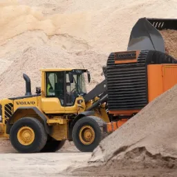 Żółta ładowarka Volvo L70F nabiera jasne trociny z pryzmy, widoczne opony z żółtymi felgami i operator w kabinie, w tle góra trocin.