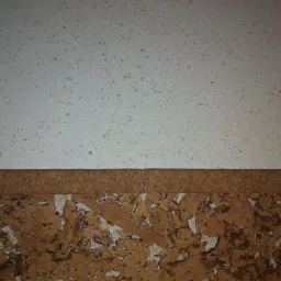 Fragment ściany z górną częścią pomalowaną na biało z brązowymi plamkami, oddzieloną korkowym paskiem od dolnej części wykończonej płytami korkowymi z widocznymi białymi fragmentami.