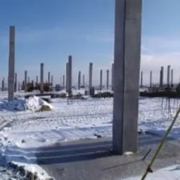 Panorama placu budowy w zimowej scenerii z widocznymi betonowymi słupami i pracownikiem w kamizelce odblaskowej obsługującym niwelator.