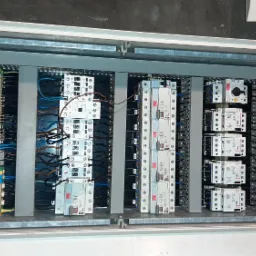 Otwarta skrzynka elektryczna z widocznymi bezpiecznikami, przewodami i złączkami, zamocowanymi na szynach DIN, z oznaczeniami na komponentach.