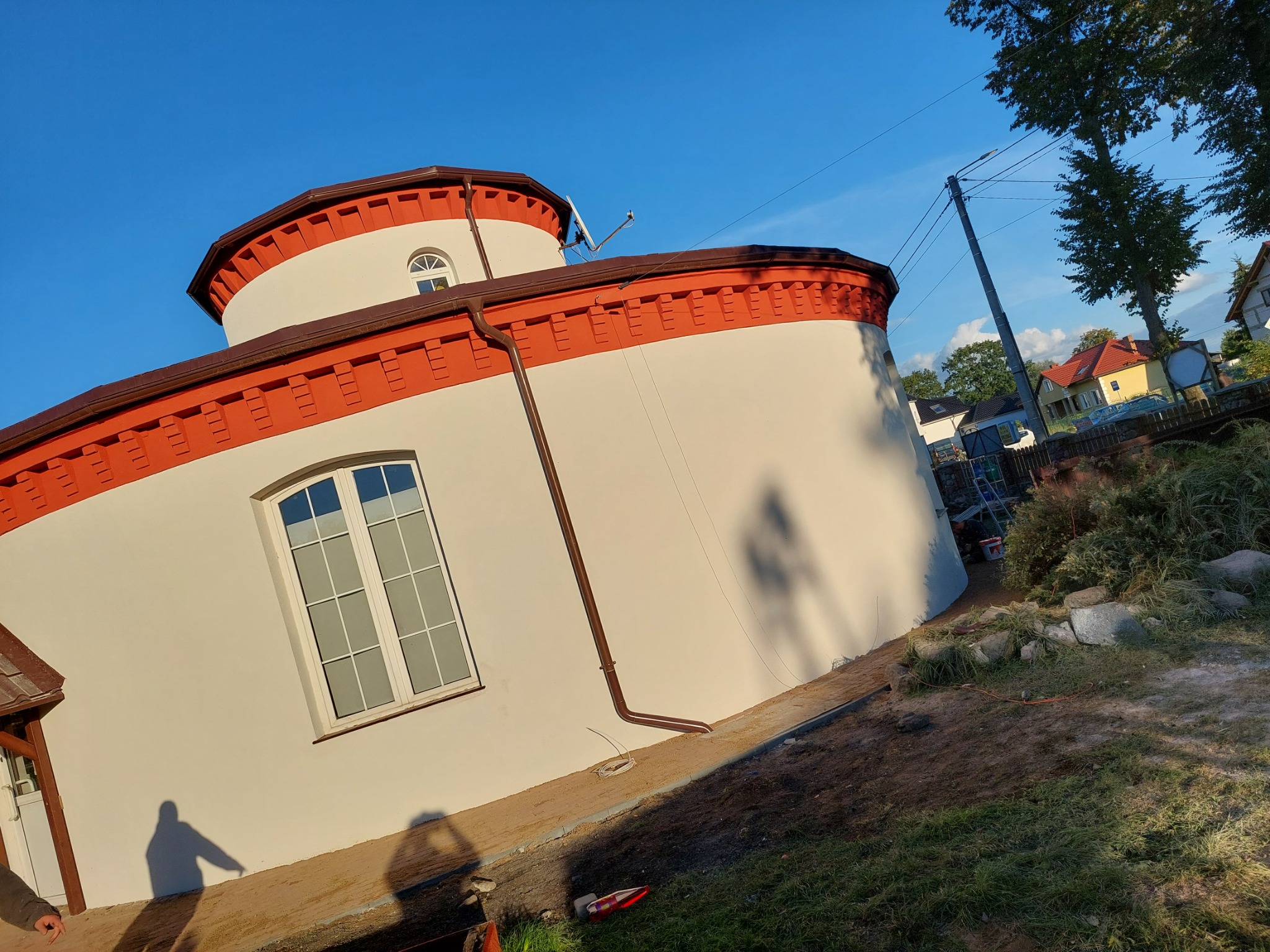 Okrągły budynek z białą elewacją i ceglastoczerwonymi detalami pod błękitnym niebem, widoczny cień osoby i częściowo zagospodarowany teren wokół.