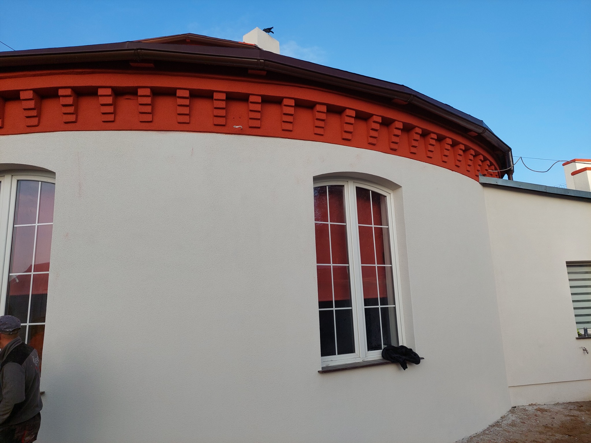 Okrągły budynek z oknami po remoncie elewacji, z dekoracyjnym, pomarańczowym pasem pod dachem i ptakiem na kominie.