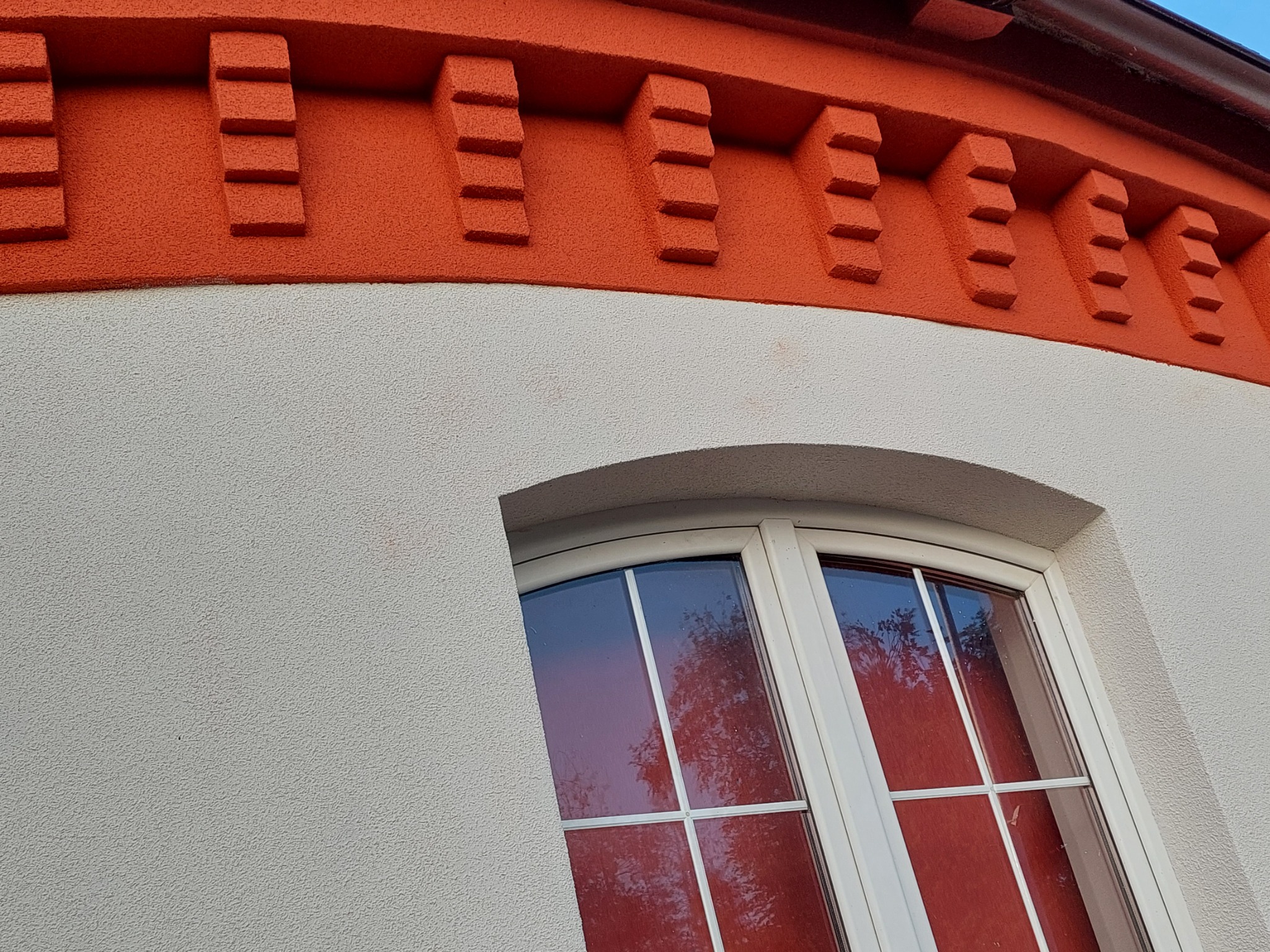 Fragment elewacji budynku z oknem łukowym, pomalowany na biało-pomarańczowo z dekoracyjnymi elementami w kolorze pomarańczowym, przypominającymi blanki.