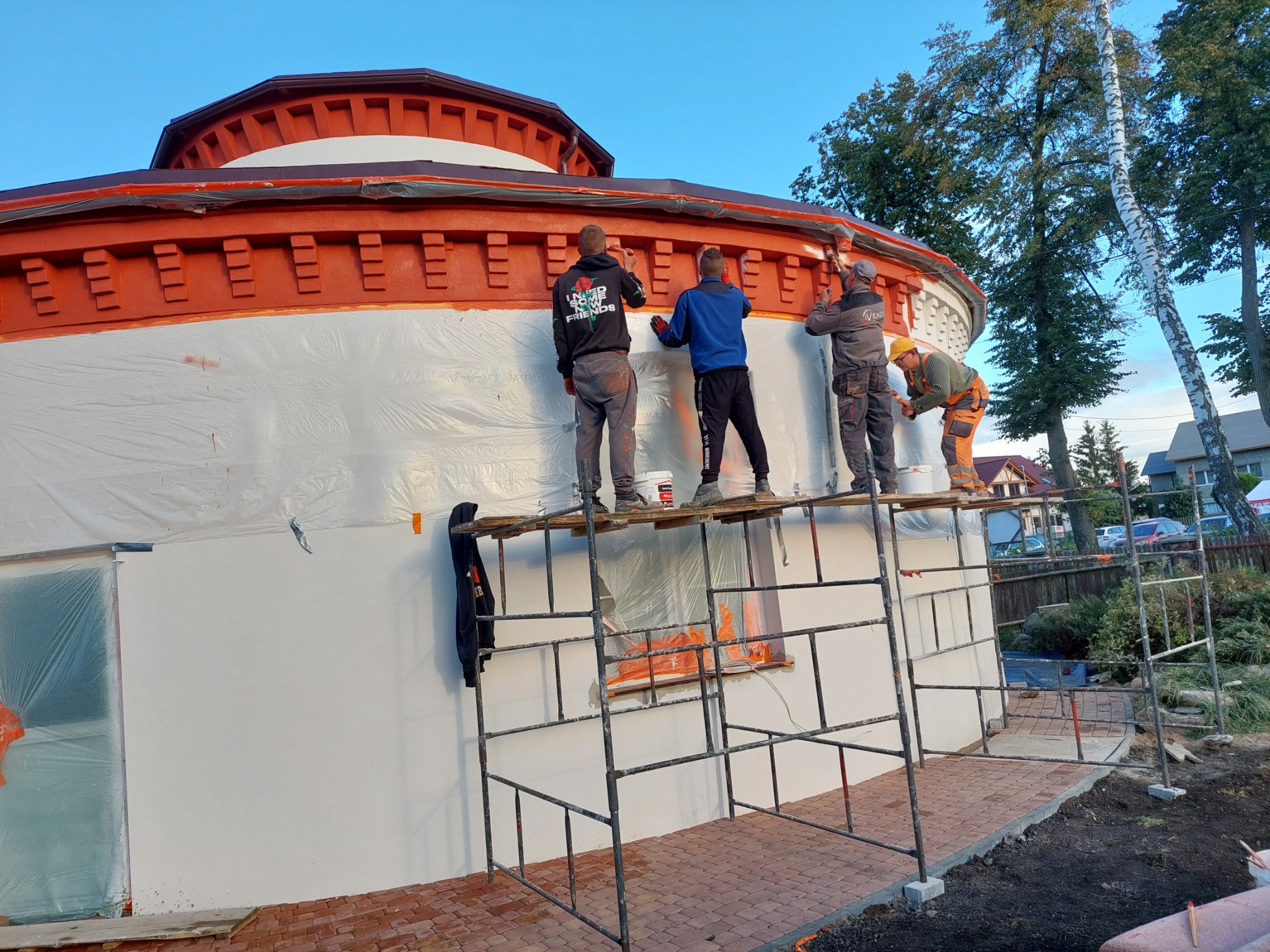 Czterech mężczyzn maluje elewację okrągłego budynku z rusztowania, zabezpieczonego folią, z widocznymi elementami dekoracyjnymi w kolorze ceglastym, na tle drzew i błękitnego nieba.