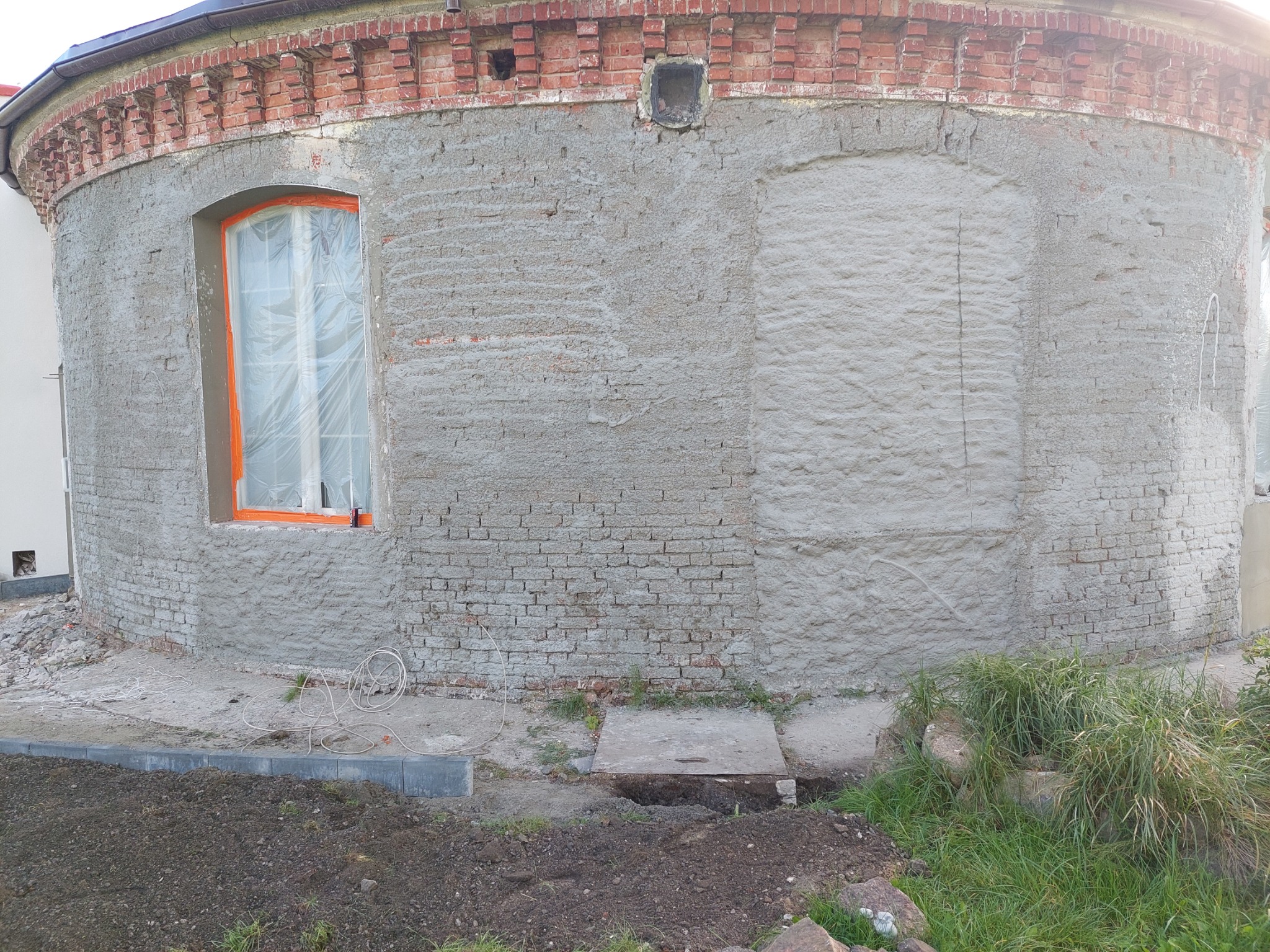 Okrągły budynek z cegły w trakcie prac elewacyjnych, z oknem zabezpieczonym folią i fragmentami świeżego tynku, widoczne ślady po zamurowanych otworach.
