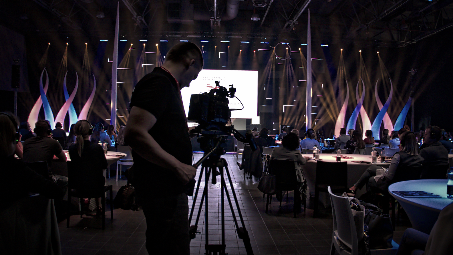 Kamerzysta w trakcie filmowania konferencji, widoczny na pierwszym planie z profesjonalną kamerą na statywie, w tle publiczność siedząca przy okrągłych stołach i oświetlona scena z dekoracjami.