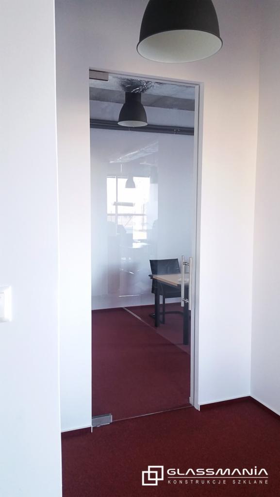 Szklane drzwi przesuwne w biurze z widocznym biurkiem i laptopem w tle, oświetlenie sufitowe.