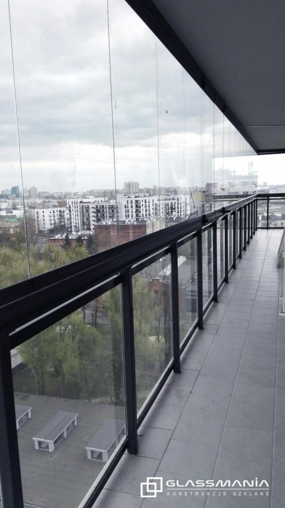Zabudowany szklanymi panelami balkon z czarną metalową balustradą, widok na miasto w tle, logo firmy w prawym dolnym rogu.