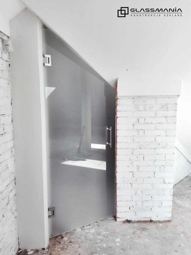 Matowa szklana ściana działowa z metalowymi okuciami, zamontowana w pomieszczeniu z białą cegłą i surowym betonem, widoczne logo firmy Glassmania.