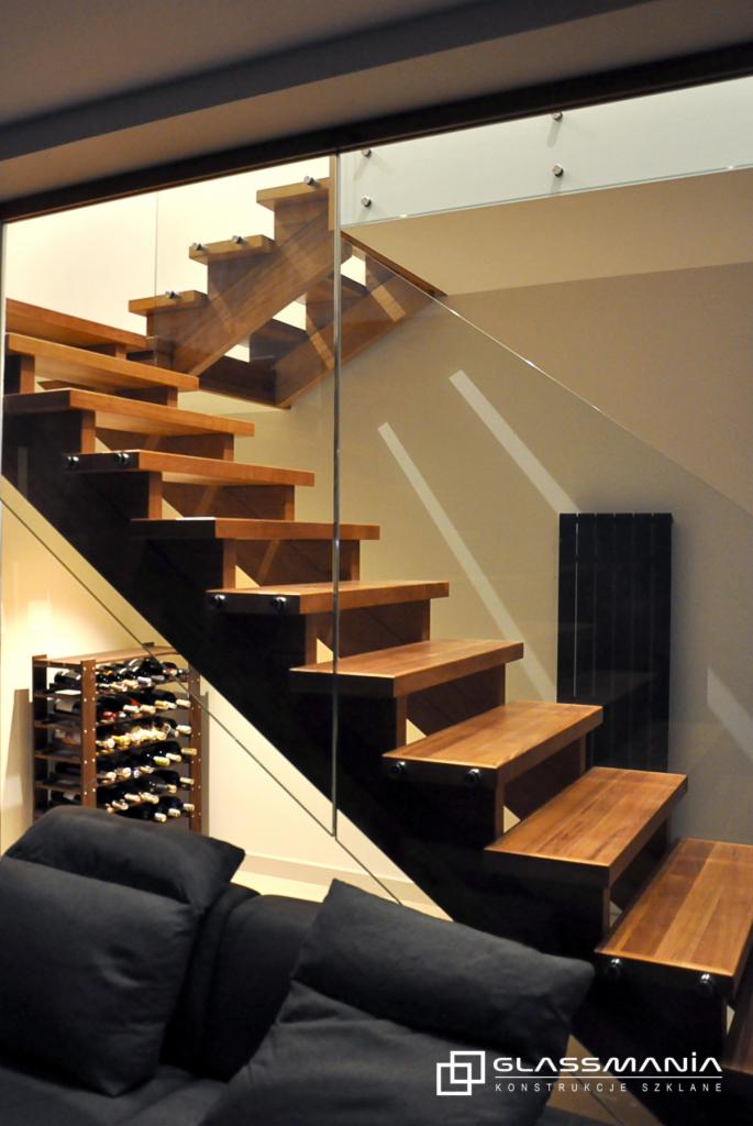 Drewniane schody z przeszkloną balustradą w nowoczesnym wnętrzu w Warszawie, widoczne regał na wino i czarna sofa.