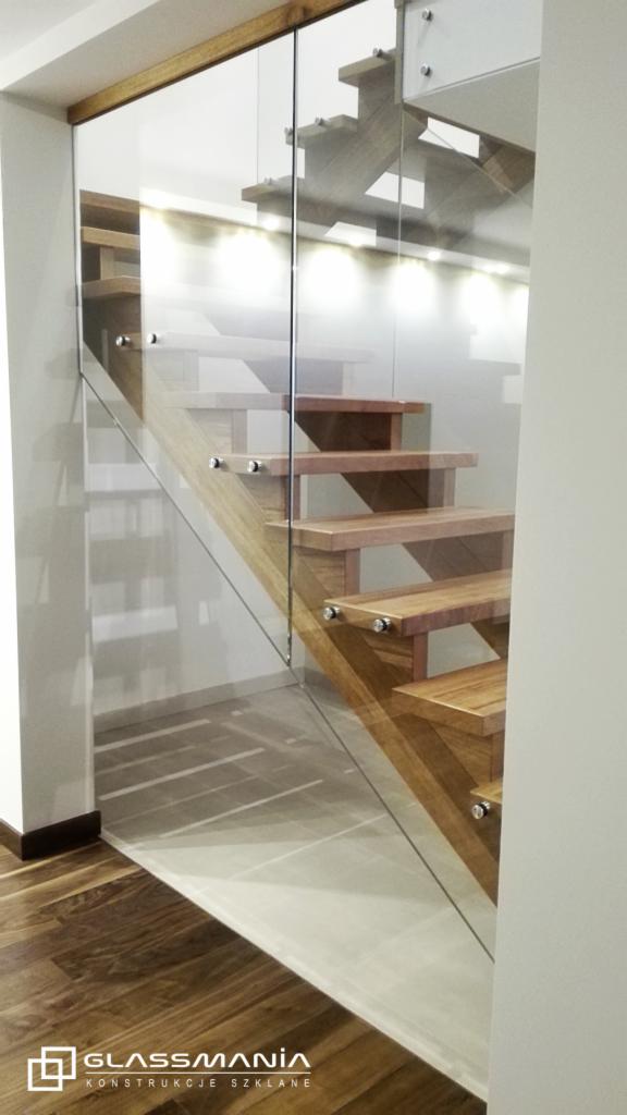 Nowoczesne schody drewniane z balustradą ze szkła hartowanego, mocowaną na metalowych uchwytach, oświetlone punktowymi lampami w ścianie.