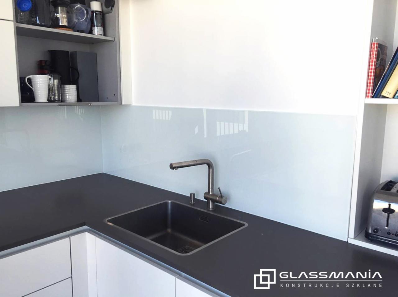 Nowoczesna kuchnia z szarym blatem, czarnym zlewem i jasnym panelem szklanym na ścianie między szafkami, widoczne logo firmy na dole po prawej.