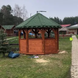 Świeżo postawiona drewniana altana ogrodowa z zielonym dachem z gontu bitumicznego na trawniku, w tle widoczne inne budynki i drzewa.