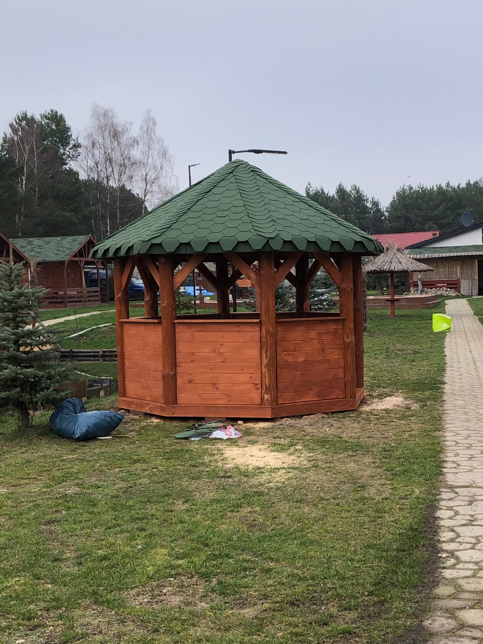 Świeżo postawiona drewniana altana ogrodowa z zielonym dachem z gontu bitumicznego na trawniku, w tle widoczne inne budynki i drzewa.