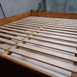 Rama łóżka z drewnianymi listwami stelaża, widoczna w surowym stanie, przed wykończeniem, w warsztacie.