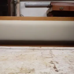 Wezgłowie tapicerowane beżową tkaniną, osadzone w drewnianej ramie, widoczne na tle warsztatu stolarskiego z narzędziami i meblami.