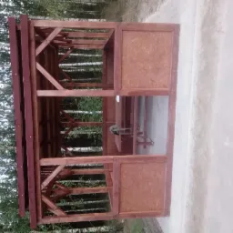 Drewniana konstrukcja altany z częściowo wypełnionymi ścianami z płyty wiórowej, widoczna na tle drzew, ujęcie z boku.