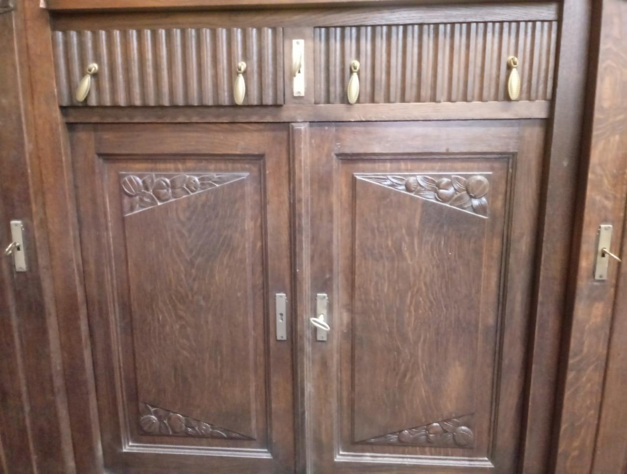 Dwudrzwiowa szafa z ciemnego drewna z rzeźbionymi motywami kwiatowymi w górnych i dolnych rogach drzwi, widoczne metalowe uchwyty i dekoracyjne pionowe żłobienia w górnej części.