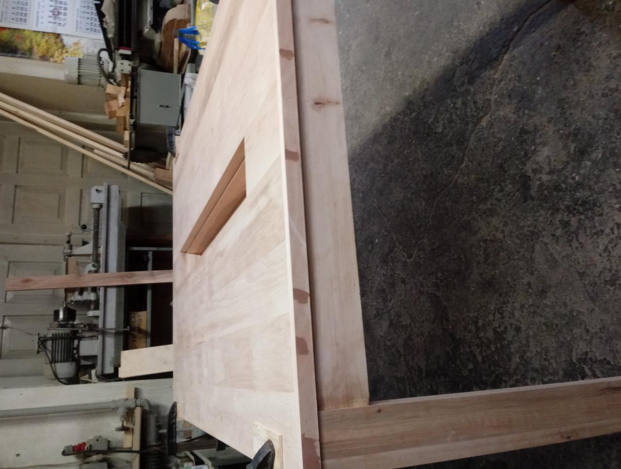 Surowe drewniane drzwi z prostokątnym otworem na szybę, ustawione pionowo w warsztacie, widoczne maszyny stolarskie i betonowa podłoga.