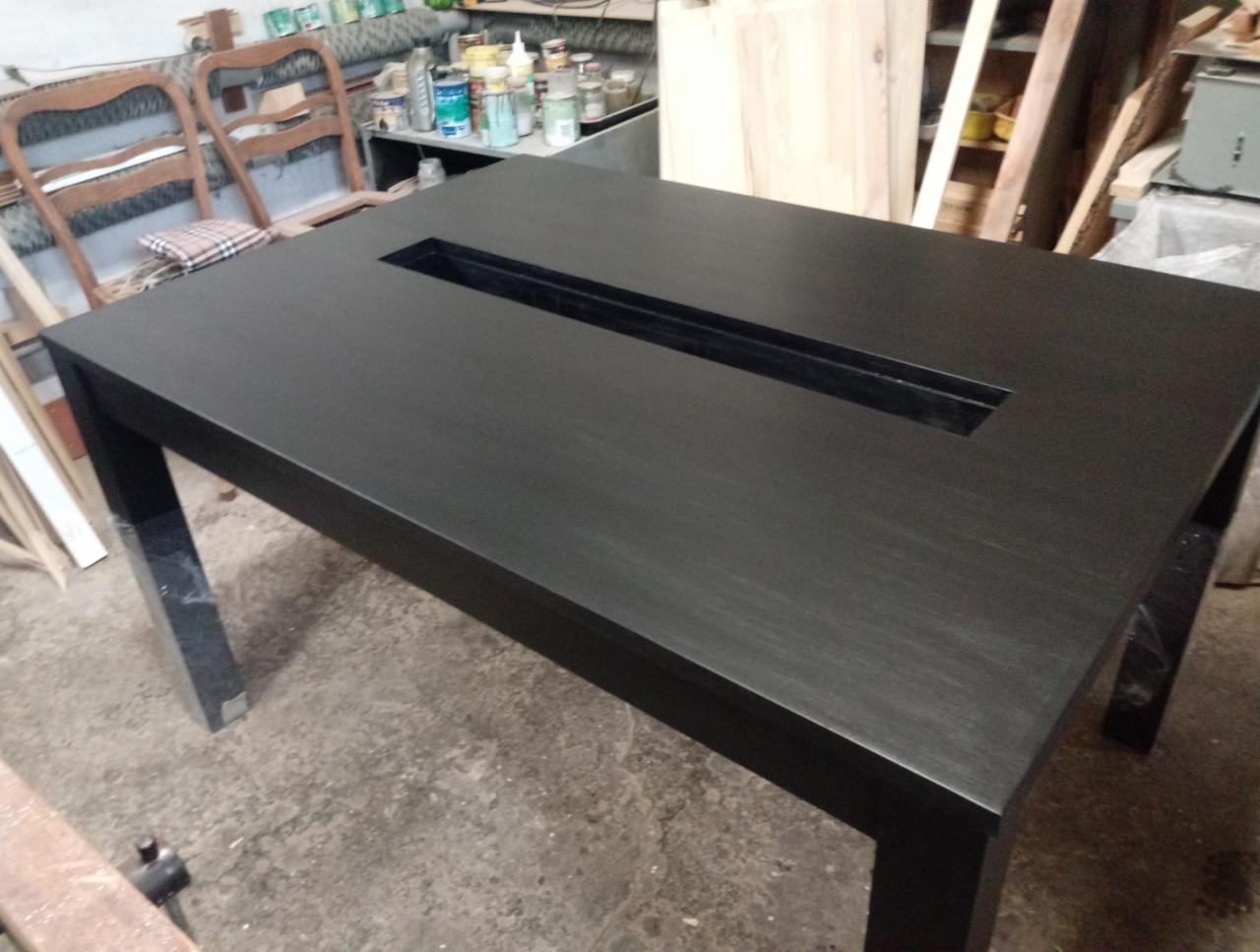 Czarny, drewniany stół z prostokątnym wgłębieniem na środku blatu, widoczny w warsztacie stolarskim z narzędziami i materiałami w tle.