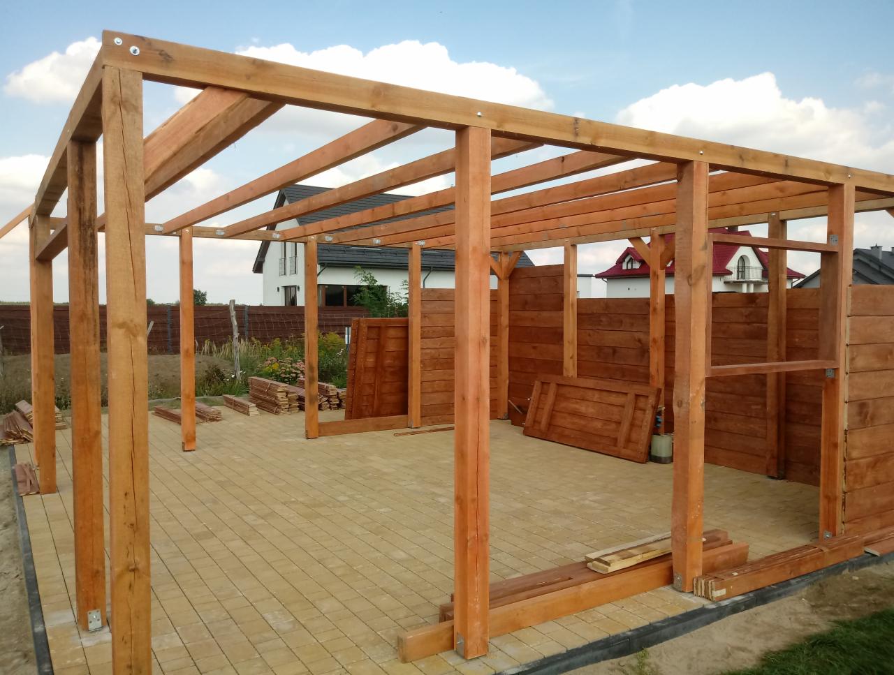 Konstrukcja drewnianej altany w trakcie budowy, widoczne słupy, legary, częściowo obite deskami ściany, kostka brukowa na podłożu, w tle domy.