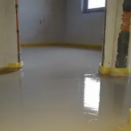 Świeżo wylana, mokra wylewka podłogowa z żółtą taśmą dylatacyjną przy ścianach i słupach nośnych, odbijająca światło okna.