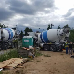 Dwa betonomieszarki z niebieskimi pasami na placu budowy domu jednorodzinnego, widoczne elementy szalunku i pracownicy w pobliżu pompy do betonu.