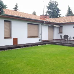 Biały dom jednorodzinny z brązowym dachem, drewnianym tarasem i zielonym trawnikiem, widok z ogrodu.